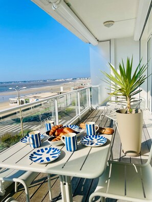 Un balcón con una mesa preparada para el desayuno, una planta en maceta y vistas a la playa y al paisaje urbano.