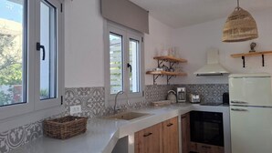 Una cocina moderna con un refrigerador blanco, un lavamanos y armarios de madera. Hay dos ventanas con persianas, una lámpara colgante y una cesta en la encimera.