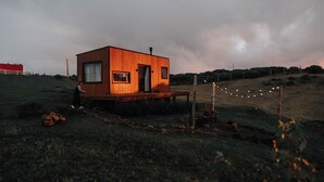 A wooden cabin with a deck, surrounded by a grassy field and a cloudy sky.
