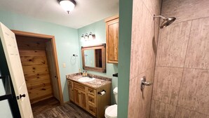A bathroom with a wooden vanity, a toilet, a shower, and a mirror with lights.