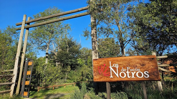 Une enseigne en bois portant l’inscription « Los de Juan Notyos », placée dans un cadre champêtre avec des arbres et un ciel bleu dégagé.