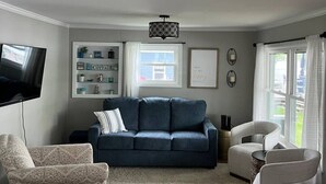 A living room with a blue sofa, patterned armchair, and white armchair. There is a flat-screen TV, a bookshelf, and a window with curtains.