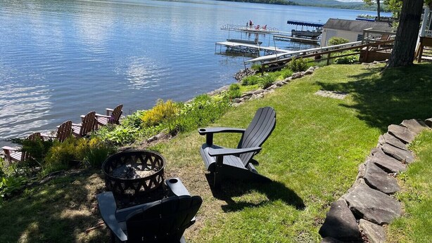 A lakeside deck with Adirondack chairs, a fire pit, and a stone pathway.