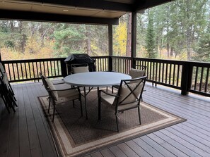 Een overdekt terras met een ronde tafel en vier stoelen, een barbecue en uitzicht op bomen.