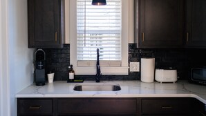 A kitchen with dark cabinets, a white countertop, a black faucet, a coffee maker, a toaster, and a window with blinds. - The Mane & Co - No. 8 (Chambersburg)