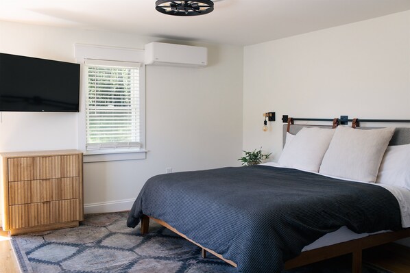 A bedroom with a bed, a TV mounted on the wall, an air conditioner, a window with blinds, a wooden cabinet, and a small plant. - The Mane & Co - No. 8 (Chambersburg)