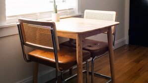 A dining area with a wooden table and two chairs, a window with blinds, and a cactus on the windowsill. - The Mane & Co - No. 8 (Chambersburg)
