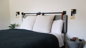 A neatly made bed with a dark green bedspread, a grey headboard, and two white pillows. There are two wall-mounted light fixtures with exposed bulbs and a small table with a remote control and a power cord. - The Mane & Co - No. 8 (Chambersburg)