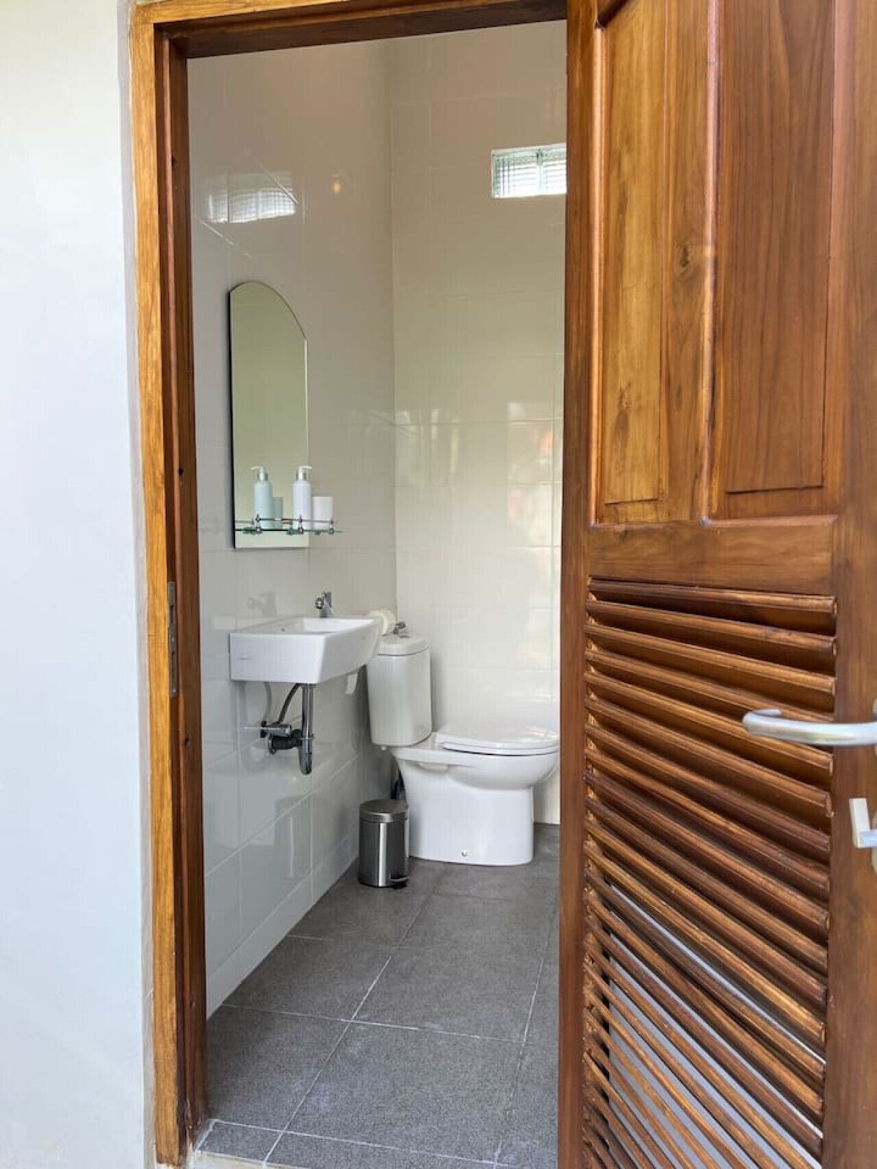 A modern bathroom with a wooden door, white tiles, a sink, a toilet, and a mirror.