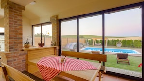 A patio with a wooden table and bench, a brick fireplace, and a large window overlooking a swimming pool and garden. - Gorgeous home in Karanac with sauna (Karanac)