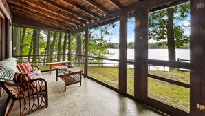 Eine überdachte Veranda mit Holzboden, einem gepolsterten Sessel, einem kleinen Tisch und großen Fenstern mit Blick auf einen See und Bäume.