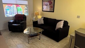 A living room with a black leather chair, a glass coffee table, a black sofa, and a small table with a lamp.