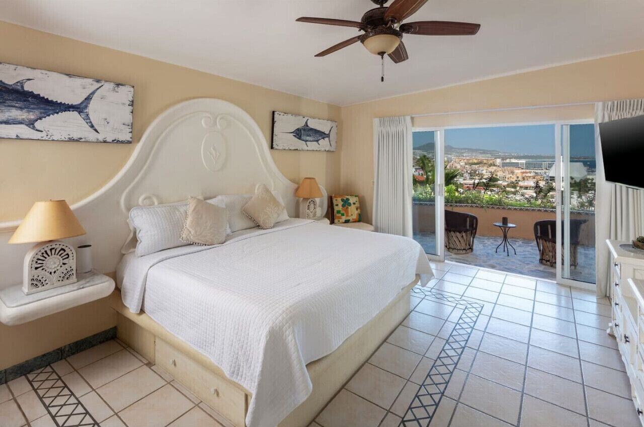 A bedroom with a large bed, a ceiling fan, and a balcony with a view of the city.