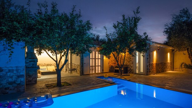 A swimming pool area with a stone wall, trees, and a building with glass doors and warm lighting. - Your private couples haven in Corfu (Halikounas)