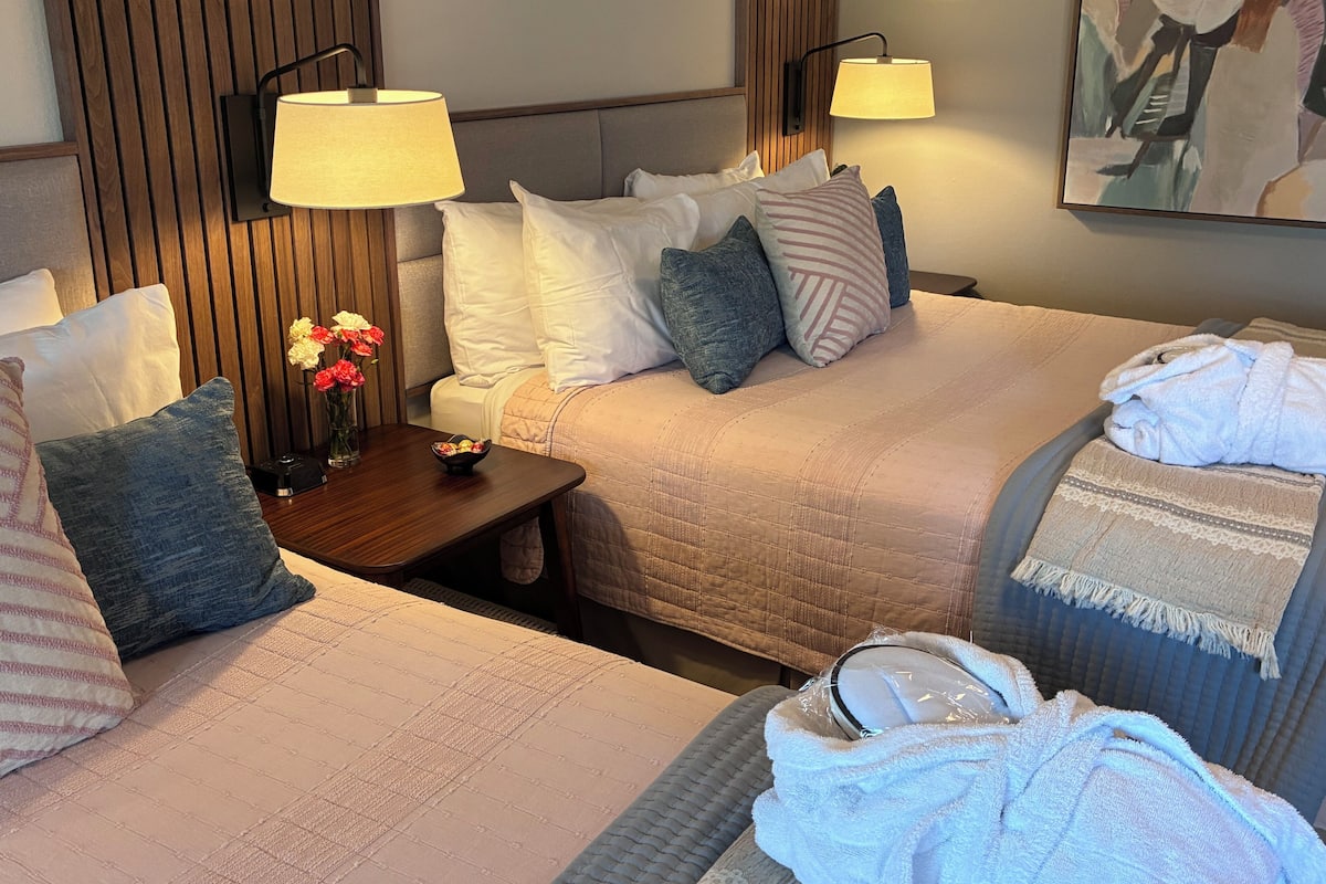A hotel room with two beds, a nightstand with a vase of flowers, two wall-mounted lamps, and a painting on the wall.