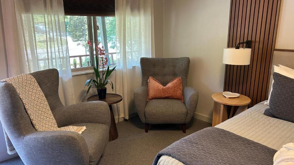 A hotel room with a bed, two armchairs, a small table, a lamp, and a window with curtains.