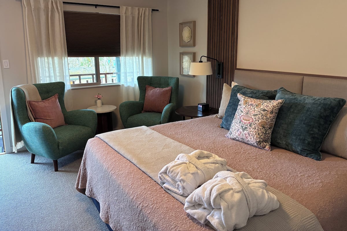 A hotel room with a bed, two armchairs, a small table, a lamp, and a window with curtains.