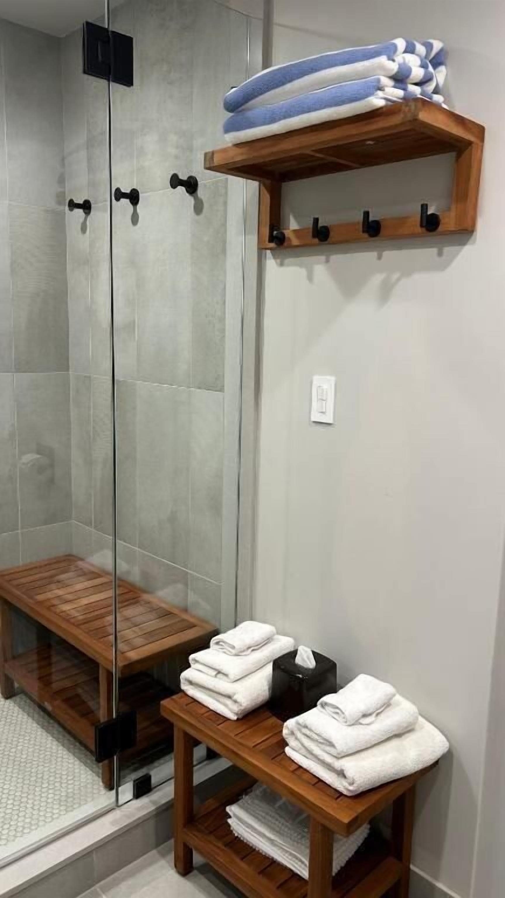 A shower area with a glass door, a wooden bench, and a shelf with towels and a tissue box.