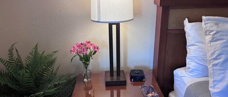 Ein Nachttisch aus Holz mit einer Lampe, einer Vase mit rosa Blumen, einer Fernbedienung und einem Blumentopf mit einem Farn.