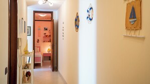 Un pasillo con una cama, un estante con varios objetos y una puerta que da a una habitación con una cama rosa y una mesita de noche.
