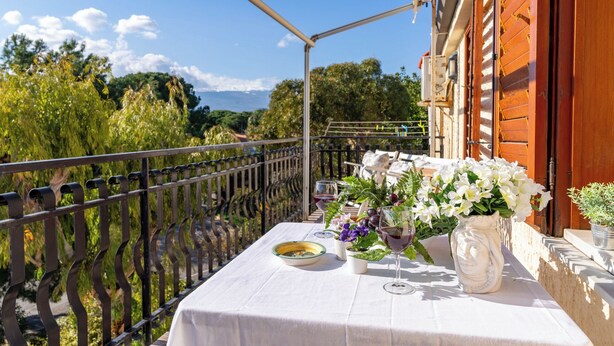 Un balcón con una mesa lista para comer, que incluye un jarrón con flores y una copa de vino.