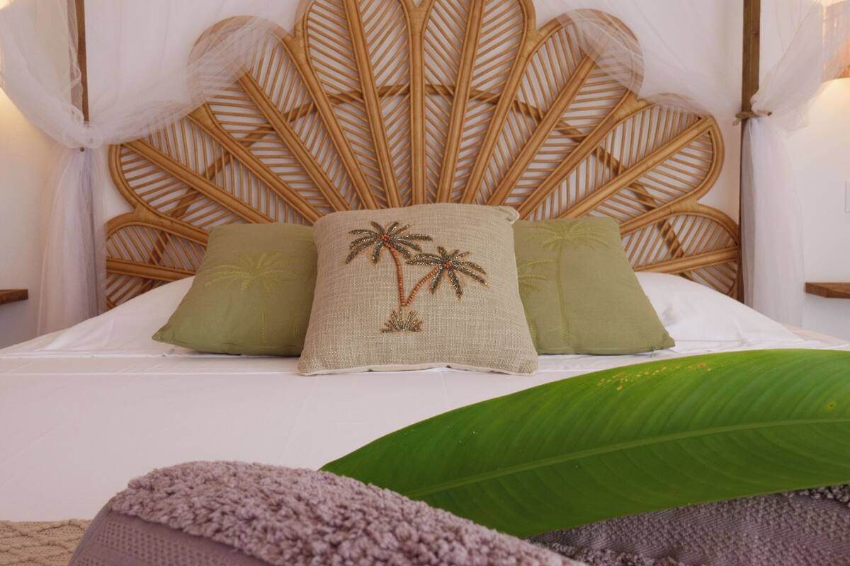 Una cama con cabecera de tejido, dos almohadas y una hoja verde grande.