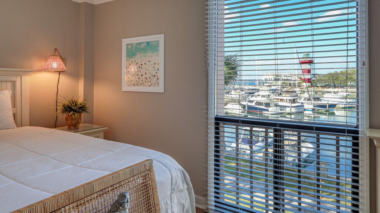 A bedroom with a bed, a nightstand, a lamp, a potted plant, and a window with a view of a marina.