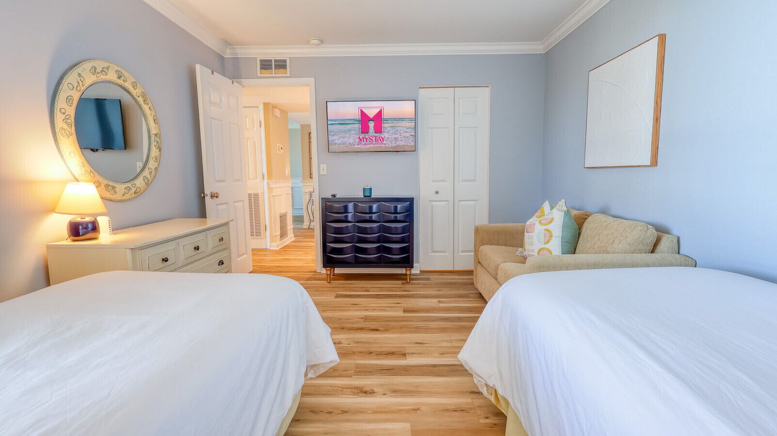 A hotel room with two beds, a dresser, a vanity with a mirror, a dresser with drawers, a sofa, and a framed picture on the wall.