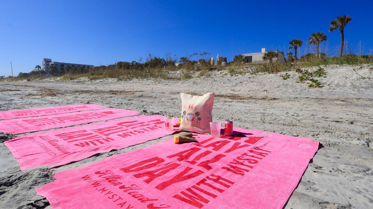 Una playa con toallas rosas extendidas sobre la arena, cada una con el texto 'ALL DAY WITH MYSTERY'.