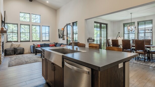 A spacious kitchen with a large island, dark wood cabinets, and a stainless steel dishwasher. There are multiple windows providing natural light and a view of the outdoors.