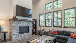 A living room with a stone fireplace, a flat-screen TV mounted above it, a gray sofa with decorative pillows, and large windows with a view of trees.