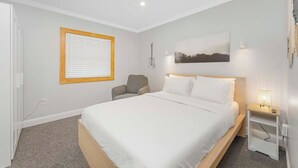 Quarto com cama, cadeira, mesa de cabeceira, guitarra presa à parede e janela com persianas.