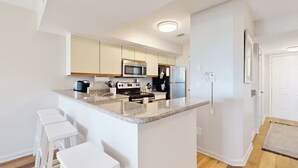 Eine moderne Küche mit weißen Schränken, einer Granit Arbeitsplatte, einer Mikrowelle, einem Herd und einem Kühlschrank. Drei weiße Barhocker und eine kleine Kochinsel mit Spüle gehören ebenfalls dazu.