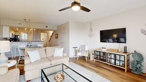 Ein modernes Wohnzimmer mit Sofa, Couchtisch, Fernseher und einem Essbereich mit Stühlen sowie einer Küche im Hintergrund.