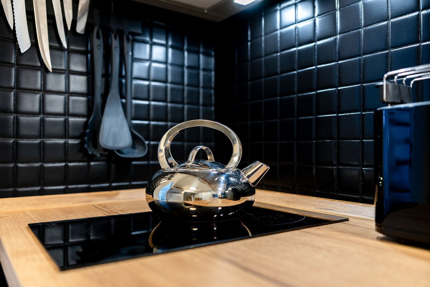 A kitchen with a stainless steel kettle on a black induction cooktop, black tiled backsplash, and a wooden countertop.