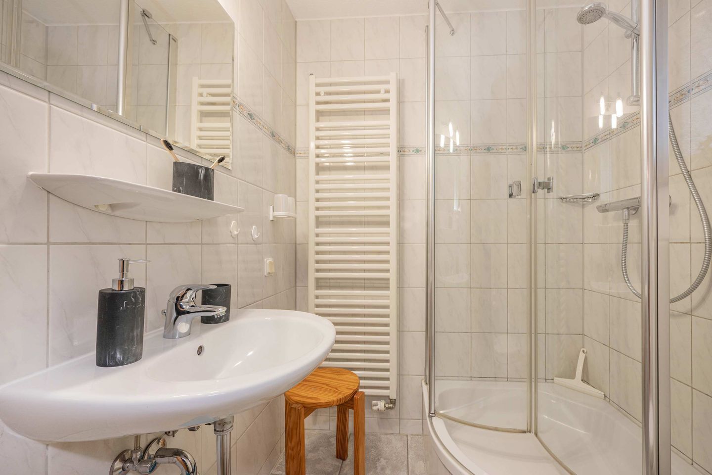 Ein modernes Badezimmer mit einem weißen Waschbecken, einem Spiegel, einem Handtuchhalter, einer Duschkabine und einem hölzernen Hocker.