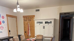 Comedor con mesa de madera, sillas estampadas, una araña y una zona de cocina con armarios blancos.