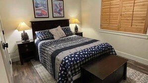 Un dormitorio con una cama, dos mesitas de noche, un banco de madera, un tapete estampado y una ventana con contraventanas de madera.