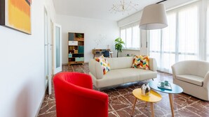 A modern living room with a sofa, red armchair, and a coffee table. There is a bookshelf, a plant, and a window with curtains. - GuestHost - This cozy and bright 124 sqm penthouse apartment can accommodate up to 6 people and is located on the 6th floor of a 1970s building with lift.The apartment boasts a splendid view over the city of Pisa, which guests can admire from one of its  (Pisa)