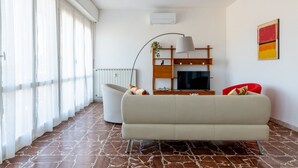 A modern living room with a sofa, a TV, a lamp, and a radiator. - GuestHost - This cozy and bright 124 sqm penthouse apartment can accommodate up to 6 people and is located on the 6th floor of a 1970s building with lift.The apartment boasts a splendid view over the city of Pisa, which guests can admire from one of its  (Pisa)