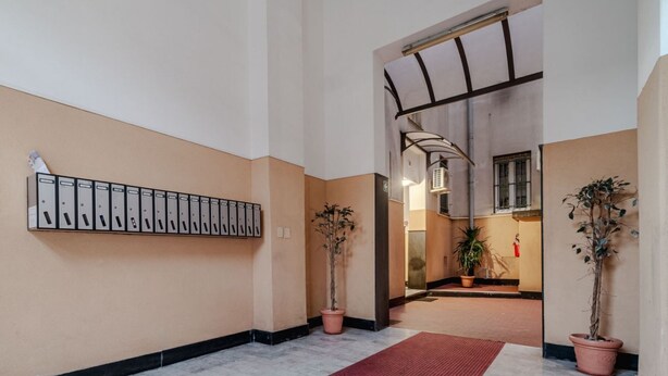 A hallway with a row of filing cabinets, potted plants, and a red carpet leading to a room with a large window and a ceiling fan. - GuestHost - Comfy APT in Genoa's St. Theodore District (Genova)