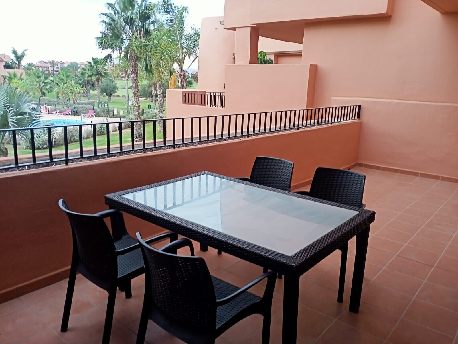 Un balcon doté d’une table à plateau en verre et de quatre chaises, offrant une vue sur une piscine et des palmiers.