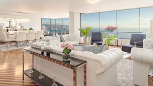 Una sala moderna con un ventanal grande que da al mar, un comedor con mobiliario blanco y una zona de estar acogedora con vistas.