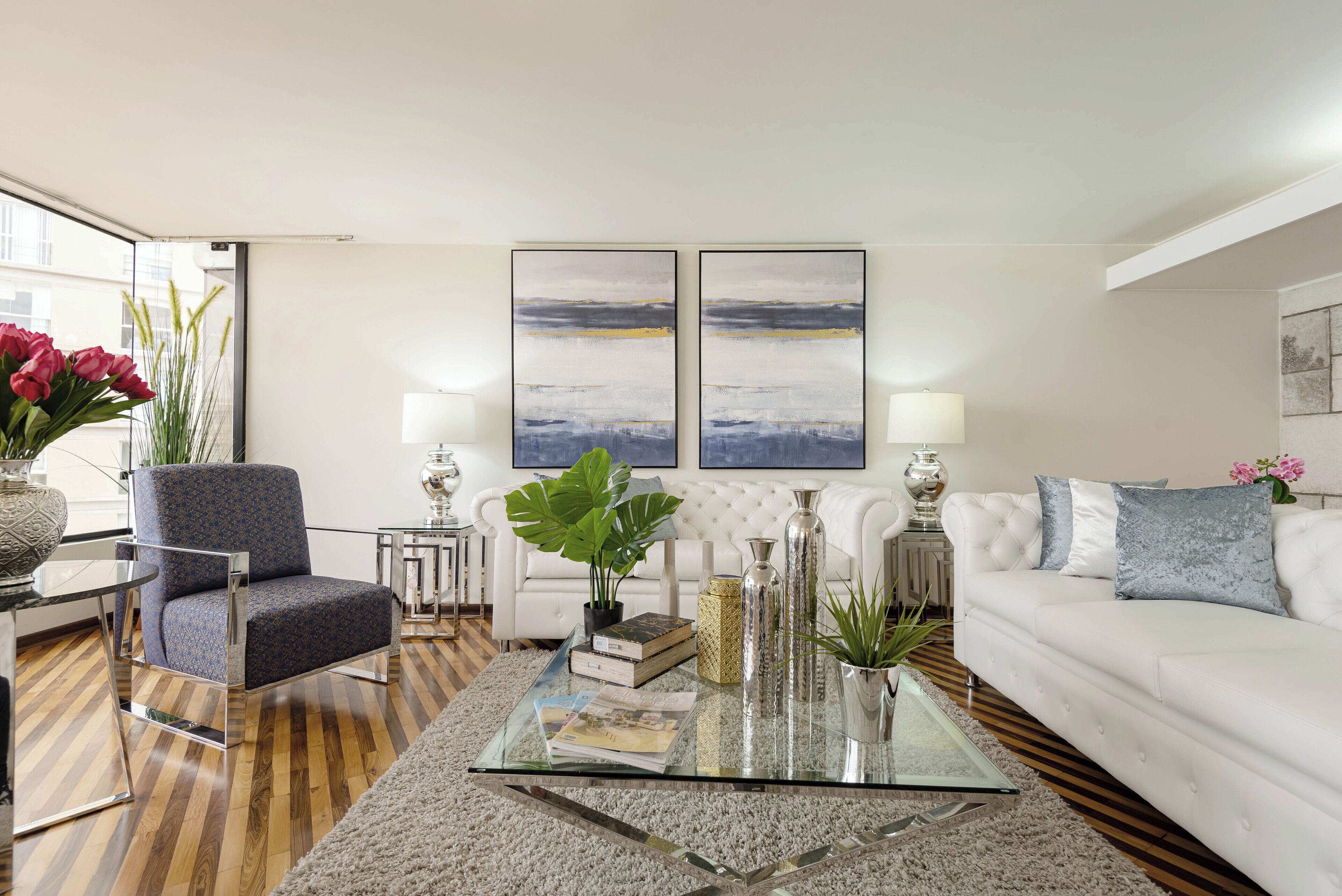 A modern living room with a glass coffee table, a white sofa, a blue armchair, and two abstract paintings on the wall.