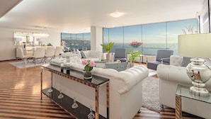 Amplia sala de estar con un gran ventanal con vista al océano, un comedor con mobiliario blanco y un diseño moderno con una mesa de centro de vidrio.