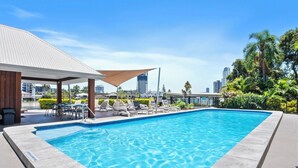 A swimming pool with lounge chairs and a shaded area, surrounded by greenery and city buildings in the background.