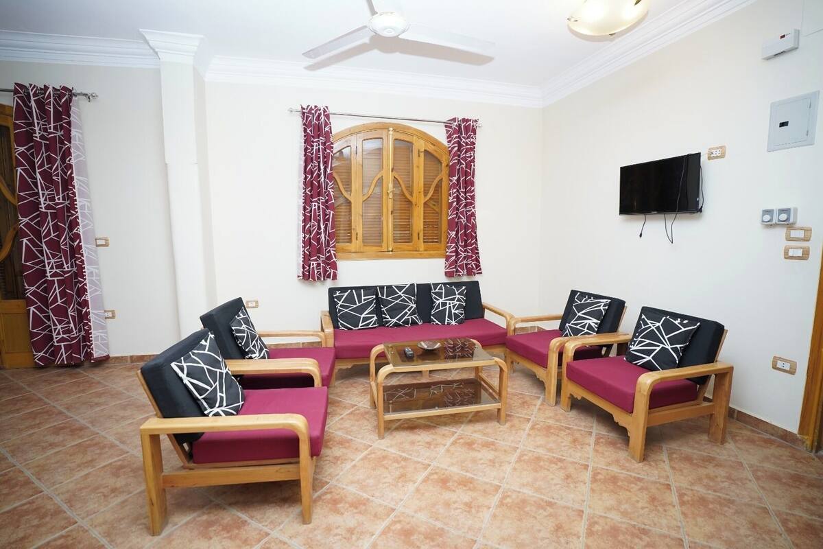 A living room with a sofa set, wooden chairs, a coffee table, a ceiling fan, a TV, and a window with shutters.