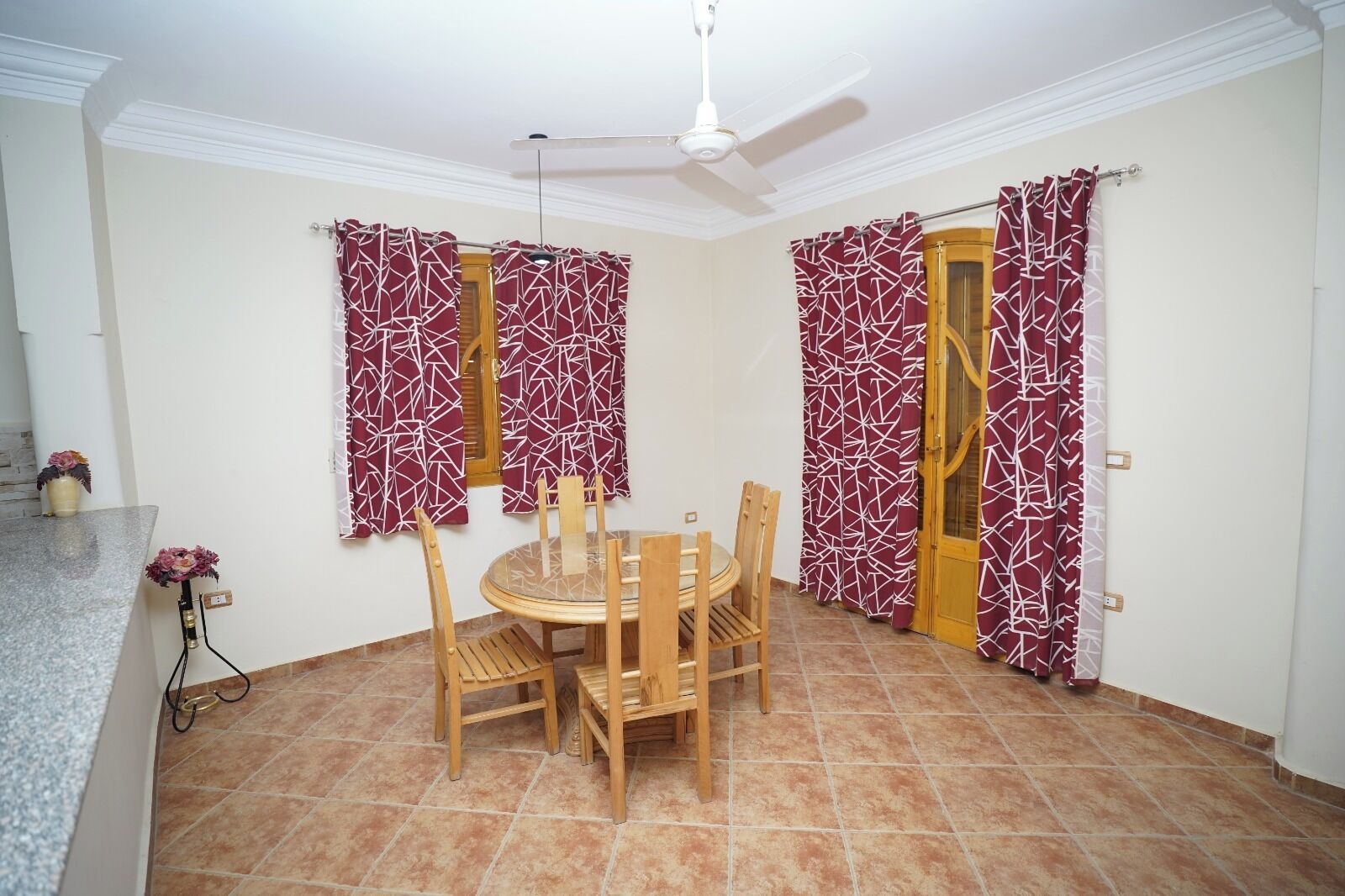 Un coin repas avec une table ronde et des chaises, deux fenêtres ornées de rideaux à motifs, un ventilateur de plafond et un sol carrelé.