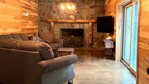 Ein gemütliches Blockhaus-Interieur mit Kamin aus Stein, einer großen Eckcouch und einem an der Wand montierten Fernseher.