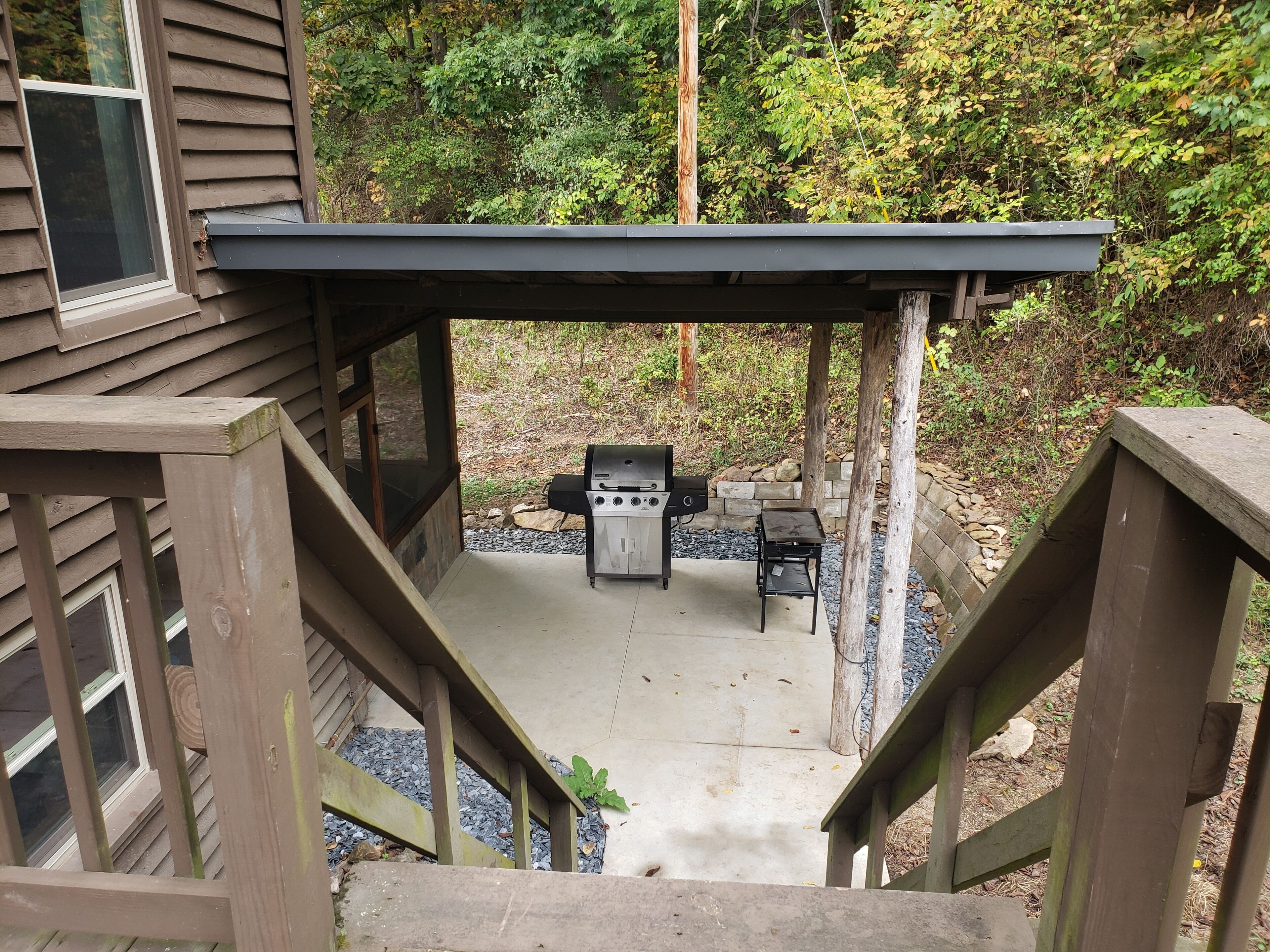 A covered outdoor patio with a grill and seating area, surrounded by wooden railings and a stone wall.